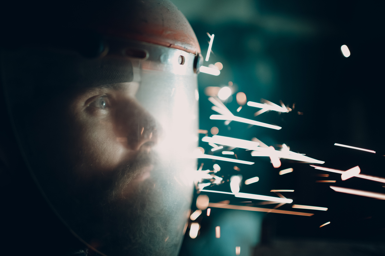 Adult bearded man in transparent protective mask and grinder saw with flying metal particles sparks in darkness