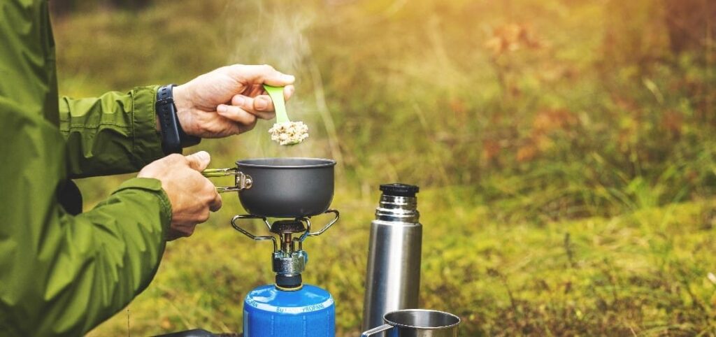 A small camping stove with someone cooking food in a small pot above it.