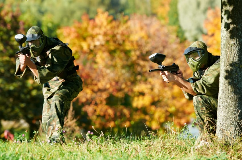 Two paintball sport players in prootective uniform and mask aiming and shoting with gun outdoors